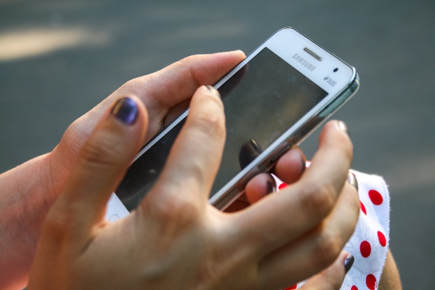 Hands holding a white Samsung smartphone with the index finger poised to touch the screen. The person, wearing a white garment with red polka dots, appears ready to use the Pocket Salon app, making appointment scheduling easier and safer.