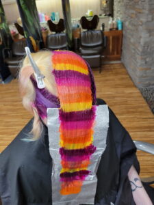 Person in a salon chair with a section of hair dyed in wide orange, yellow, and magenta stripes; foil and hair clip are used for the coloring process. - Varuna Salon Spa in Annapolis, MD