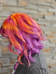Person with wavy hair dyed in bright orange, red, and purple shades, standing sideways against a blurred stone wall background. - Varuna Salon Spa in Annapolis, MD