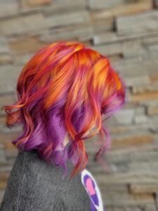 Person with short, wavy hair dyed in vibrant orange, red, and purple shades, standing in front of a stone wall background. - Varuna Salon Spa in Annapolis, MD