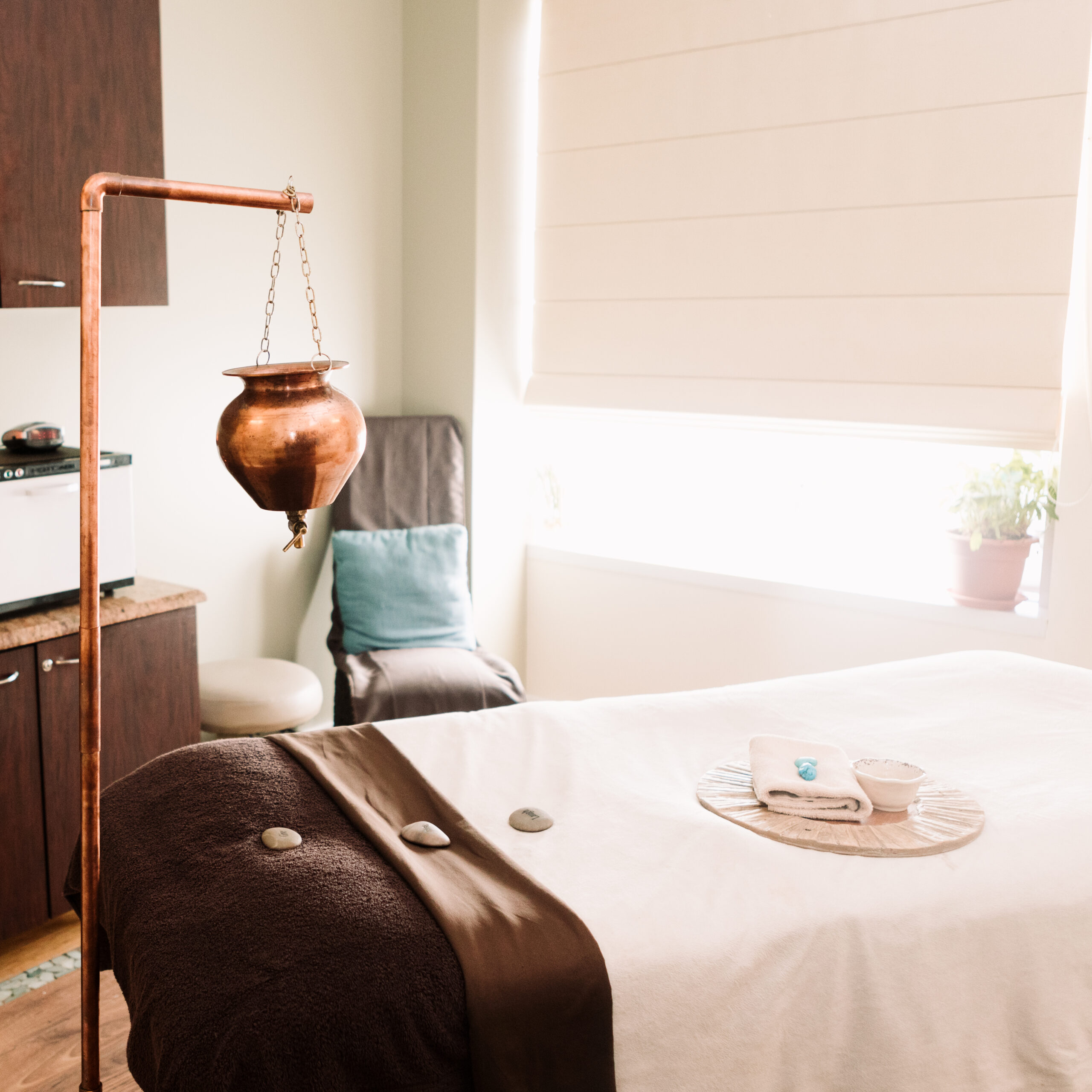 A serene spa room with a massage table, stones, a towel, and a cup. A copper pot hangs nearby, and light pours in from a window with a plant on the sill.