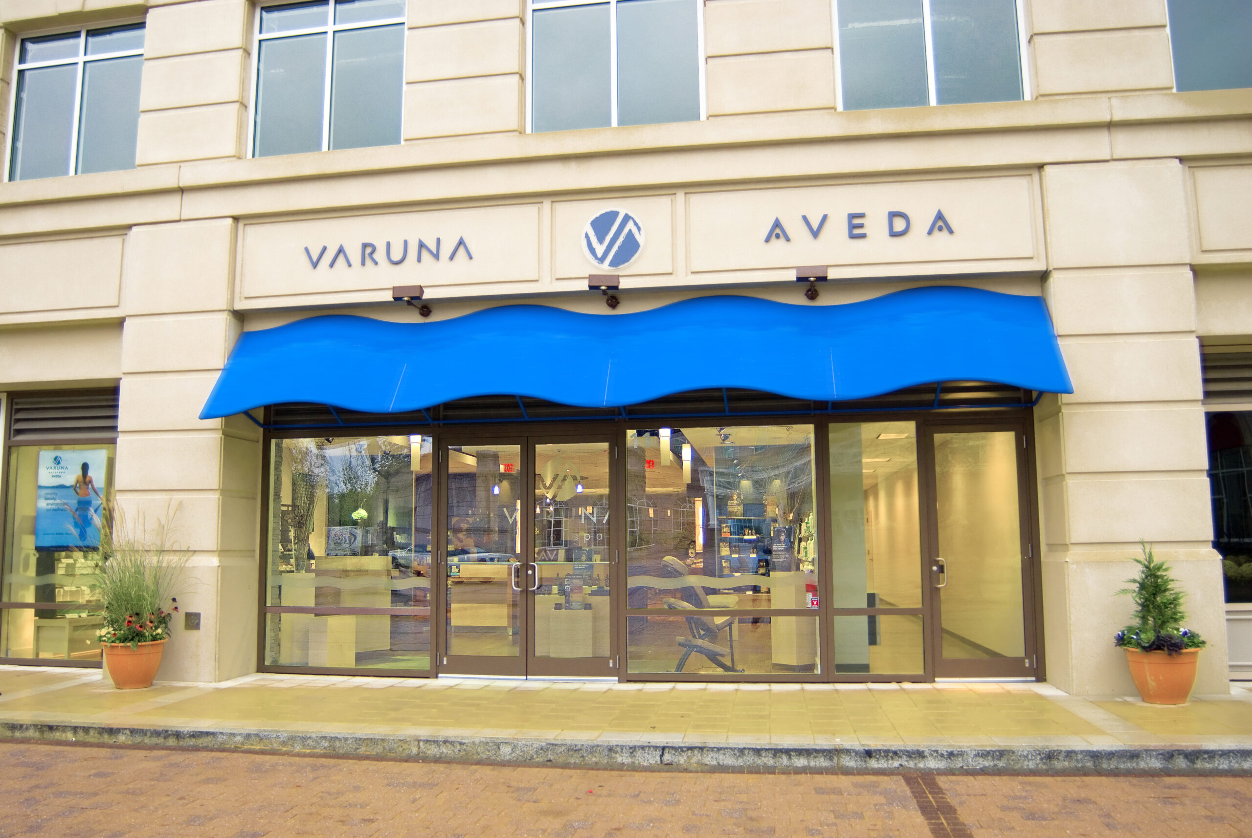 The exterior view of the building features a blue awning with "Varuna Aveda" proudly displayed above the glass doors, hinting at beautiful beginnings. The welcoming entrance, flanked by potted plants, promises a serene escape for all who enter.