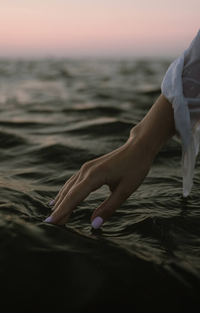A person with a white sleeve gently touches the surface of dark water at sunset, evoking the tranquility found after spa treatments Annapolis MD offers. - Varuna Salon Spa in Annapolis, MD