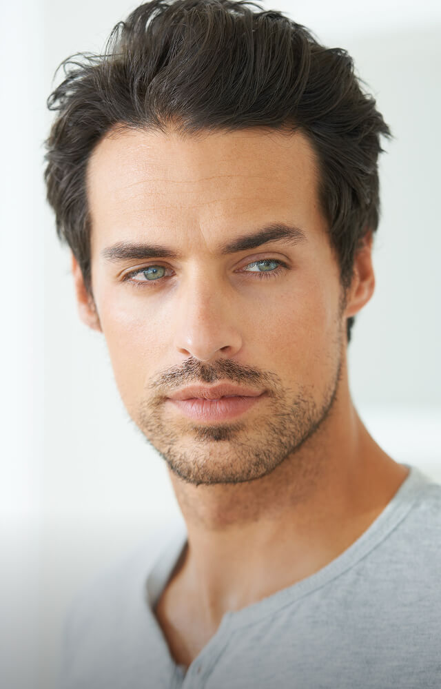 A man with short dark hair, light eyes, and light facial hair looks to the side while wearing a light gray shirt at a hair salon downtown Annapolis. - Varuna Salon Spa in Annapolis, MD