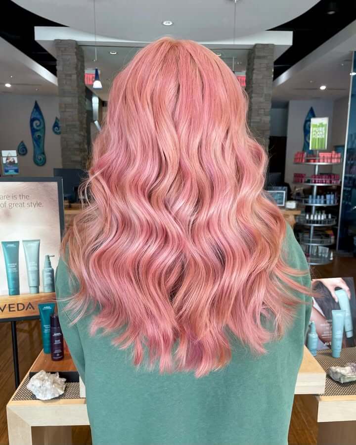 Person with long, wavy pastel pink hair stands facing away in the best salon Annapolis MD, wearing a green top. Haircare products and chic salon decor are visible in the background. - Varuna Salon Spa in Annapolis, MD