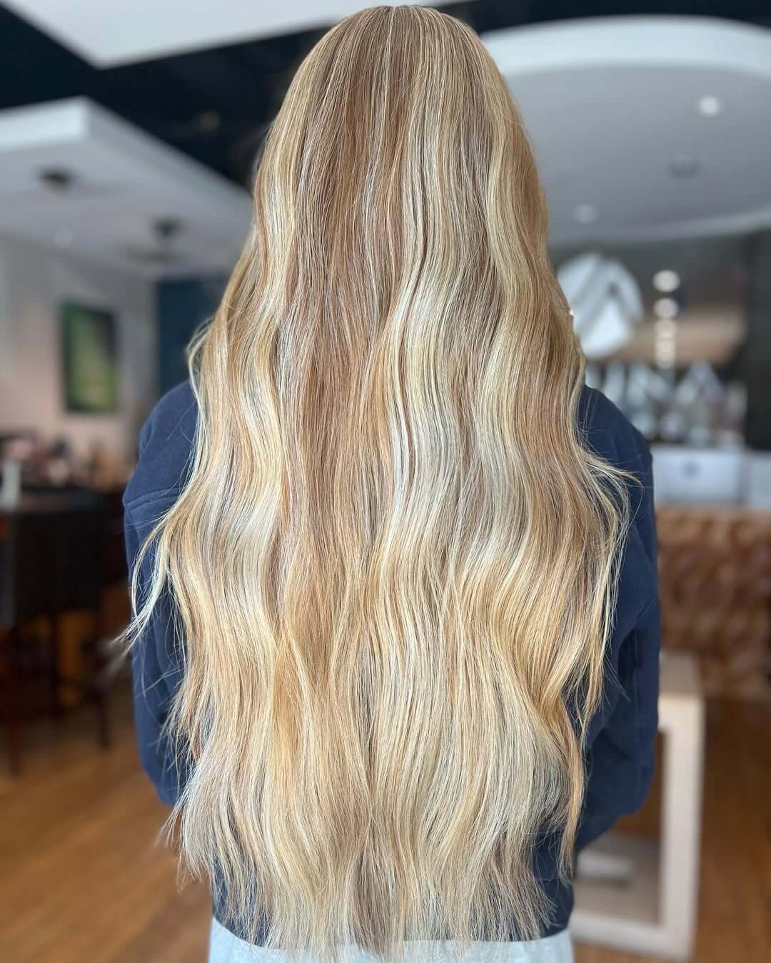 Person with long, wavy, blonde hair seen from behind, standing indoors in a well-lit room with blurred background—perfect inspiration for those seeking professional waxing services Annapolis or rejuvenating spa treatments Annapolis MD. - Varuna Salon Spa in Annapolis, MD