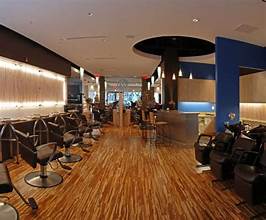 Modern hair salon interior with empty styling chairs, wooden flooring, and ambient lighting. The reception desk is visible in the background. - Varuna Salon Spa in Annapolis, MD