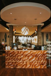 A modern spa reception area with a carved wooden desk, product shelves, and a sign reading "Varuna Salon Spa + Spa" on the back wall. - Varuna Salon Spa in Annapolis, MD