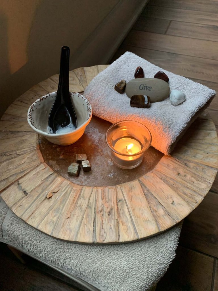 A wooden tray holds a lit candle, a bowl with a black spoon, a few crystals, and a folded towel with stones, including one engraved with "Give. - Varuna Salon Spa in Annapolis, MD