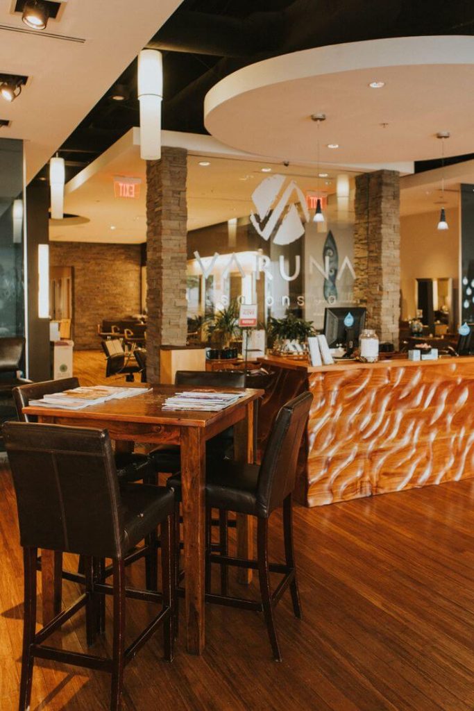 Modern salon interior with wooden table and chairs in the foreground, a wavy-patterned reception desk, and salon stations visible in the background. - Varuna Salon Spa in Annapolis, MD