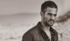 A man with short dark hair and stubble stands outdoors, looking at the camera. He wears a button-up shirt. The blurred, sandy landscape behind him suggests a moment of relaxation—perfect inspiration for Father's Day gifts like a massage or head spa. - Varuna Salon Spa in Annapolis, MD