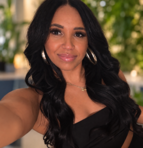 Brittany Roby, a woman with long dark hair, wearing hoop earrings and a black top, takes a selfie indoors with a blurred green background. - Varuna Salon Spa in Annapolis, MD